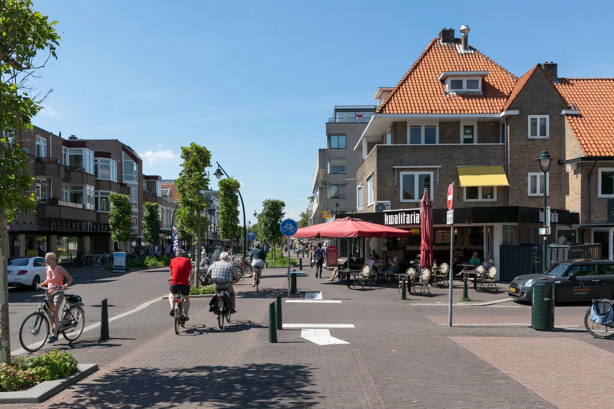 Fietsers en voetgangers bewegen zich door de zonnige winkelstraat 1e Hogeweg met terras en winkels, waaronder Kwalitaria.
